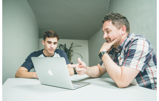 A consultant aiding a client by showing him documents on his computer as they sit at a table.